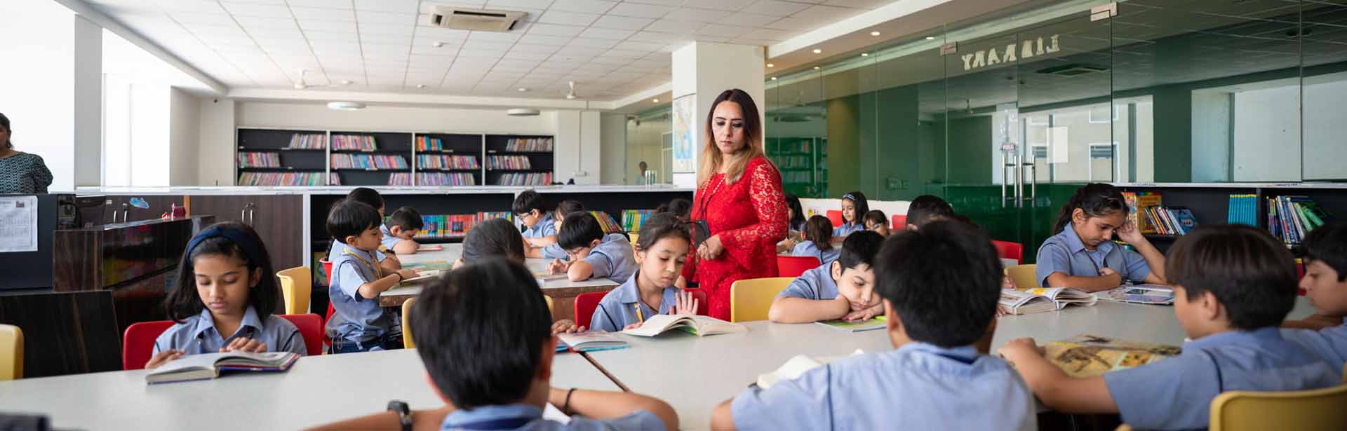 Teacher in red supervises reading at a library, showcasing the best CBSE school in Gurgaon.